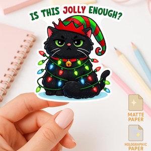 May include: A black cat sticker with a Christmas elf hat and string lights. The sticker has the text "IS THIS JOLLY ENOUGH?" in green and white. The sticker is held by a hand.