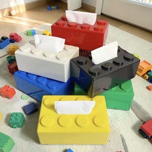 Peut inclure: Plusieurs boîtes à mouchoirs colorées en forme de grosses briques de construction. Les boîtes sont rouges, blanches, noires, bleues, vertes et jaunes, avec des mouchoirs blancs qui dépassent. Des voitures et des blocs de construction sont éparpillés.