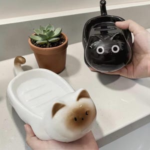 Op de afbeelding: Twee keramische katvormige zeepbakjes, één wit met bruine accenten en de andere zwart. Er is ook een kleine vetplant in een pot te zien. De kattenbakjes hebben een staart en een schaalachtig gedeelte om zeep in te bewaren.
