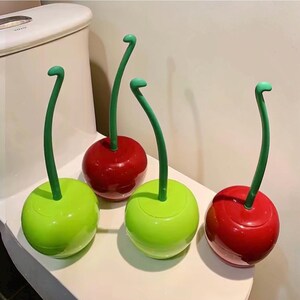 Peut inclure: Quatre brosses de toilette en forme de cerise. Deux sont rouges et deux sont vertes. Chaque brosse a un long manche vert incurvé. Les brosses sont posées sur une surface blanche, probablement une étagère de salle de bain ou le dessus des toilettes.