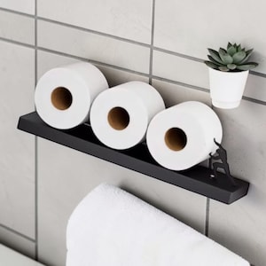 Op de afbeelding: Zwart metalen plankje met drie rollen wit toiletpapier. Rechts staat een kleine witte pot met een vetplant. Een witte handdoek hangt eronder. Het plankje heeft een decoratief silhouet van een klimmende persoon.