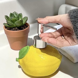 Peut inclure: Un distributeur de savon en céramique jaune en forme de citron avec une pompe argentée. Une petite plante succulente en pot est à gauche. Le distributeur a un détail de feuille verte. Le distributeur est sur une surface blanche.