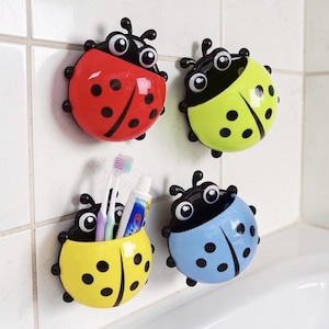 Peut inclure: Quatre porte-brosses à dents en forme de coccinelles, rouges, jaunes, verts et bleus. Chaque support a des points noirs et une tête noire avec de grands yeux blancs. Un support jaune contient des brosses à dents et du dentifrice.