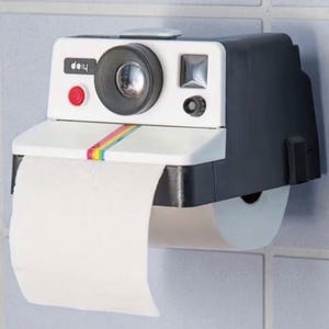 Puede incluir: Un original portarrollos de papel higiénico con forma de cámara instantánea vintage. El soporte es blanco con una sección negra, una franja arcoíris y un botón rojo. El rollo de papel higiénico es visible y dispensando.