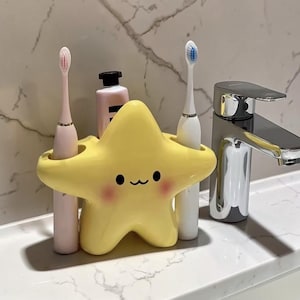 Peut inclure: Un porte-brosse à dents jaune en forme d'étoile avec un visage souriant, tenant deux brosses à dents. Le support a des yeux noirs, une bouche incurvée et des joues roses. Les brosses à dents sont roses et blanches.