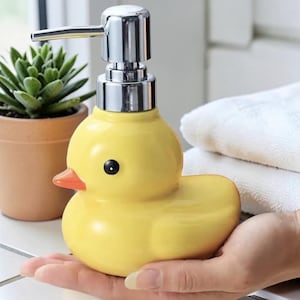 Könnte beinhalten: Ein gelber Seifenspender in Entenform aus Keramik mit einer Chrompumpe. Die Ente hat ein schwarzes Auge und einen orangefarbenen Schnabel. Eine kleine Sukkulentenpflanze in einem Terrakottatopf befindet sich im Hintergrund.