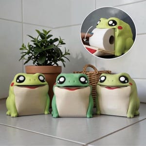 May include: Three frog-shaped toilet paper holders in shades of green and cream. The frogs have large, cartoonish eyes and red accents. One holder is open, revealing a roll of toilet paper. A small plant in a terracotta pot and a wicker basket are in the background.
