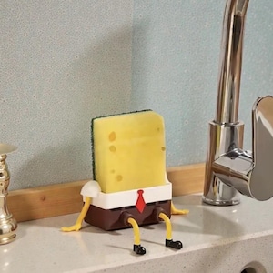 May include: A yellow sponge with a green scrubbing surface, designed to resemble a cartoon character, sits on a countertop. The sponge has a brown base with yellow legs and black shoes, a white shirt, and a red tie.