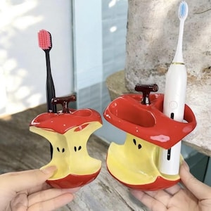 Könnte beinhalten: Zwei Zahnbürstenhalter aus Keramik in Form von Apfelhälften. Die Halter sind rot mit gelben Innenräumen und braunen Stielen. Einer hält eine schwarze Zahnbürste mit rosa Kopf, der andere eine weiße elektrische Zahnbürste.