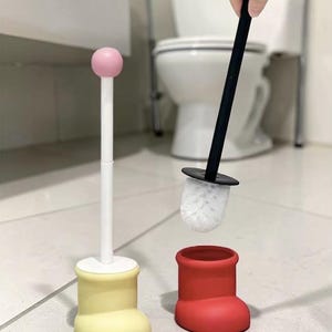 Peut inclure: Un ensemble de brosses de toilette fantaisistes. L'ensemble comprend une base jaune en forme de botte et un support surmonté d'un rose. La brosse noire a une tête en poils blancs. L'ensemble est conçu pour ajouter une touche amusante à la décoration de la salle de bain.