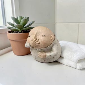 Peut inclure: Une sculpture en céramique fantaisiste représentant une figure avec une grosse tête et un petit corps, se couvrant la bouche avec sa main. La figurine porte un pull clair. Une petite plante succulente dans un pot en terre cuite est à gauche.