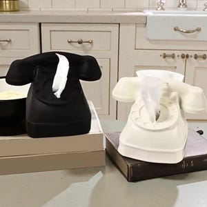 May include: Two telephone-shaped tissue box holders. One is black, the other is white. Both have a classic rotary phone design with a tissue emerging from the mouthpiece. They sit on a table with a neutral background.