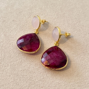 Peut inclure: Une paire de boucles d'oreilles à clous dorées. Chaque boucle d'oreille présente une pierre précieuse en forme de goutte d'eau, rouge foncé et facettée, sous une pierre ovale rose pâle plus petite. Les boucles d'oreilles sont posées sur un fond neutre.
