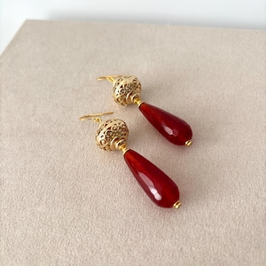 Può includere: Un paio di orecchini pendenti con pietre rosso scuro sfaccettate a forma di goccia, sospese a perline metalliche dorate sferiche e decorate con intricati motivi in filigrana e ganci abbinati per un aspetto elegante e raffinato.
