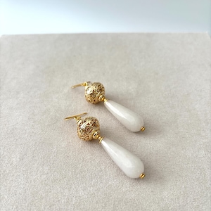 Può includere: Un paio di orecchini pendenti con pietre bianche lisce a forma di goccia sospese da perline sferiche in metallo dorato con intricati motivi in filigrana. Gli orecchini includono ganci dorati per il fissaggio.
