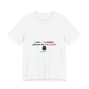Può includere: T-shirt bianca con la scritta "i am 6.7 a stable please do not patch" in nero, verde e rosa. Un personaggio dei cartoni animati con una barra di caricamento è sotto. Girocollo.