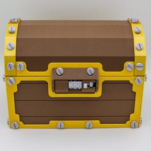 Puede incluir: Un cofre del tesoro marrón con bordes amarillos y tornillos plateados. El cofre tiene una cerradura de combinación y está hecho de un material similar a la madera. El cofre está cerrado y descansa sobre una superficie blanca.