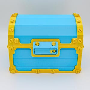 May include: A blue treasure chest with gold-colored trim and hardware. The chest has a rectangular shape with a curved top and a latch on the front. The gold trim is detailed with simulated screw heads.