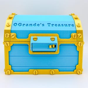 May include: A blue treasure chest with gold-colored accents and hardware. The text "O'Grande's Treasure" is embossed on the lid. The chest has a rectangular shape with a curved top and a latch closure.