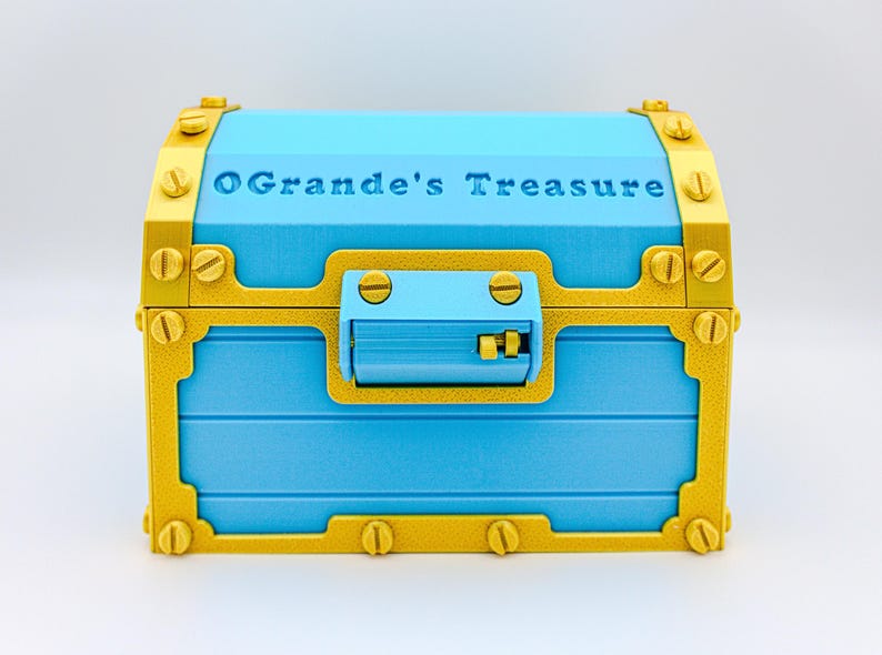 May include: A blue treasure chest with gold-colored accents and hardware. The text "O'Grande's Treasure" is embossed on the lid. The chest has a rectangular shape with a curved top and a latch closure.