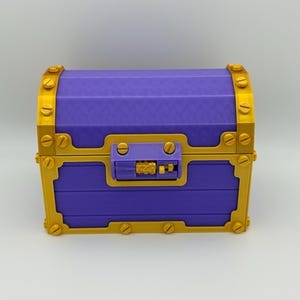 May include: A purple treasure chest with gold-colored accents. The chest features a rectangular shape with a curved top, gold hinges, and a gold clasp. The chest is made of a ridged material and has a lock on the front.