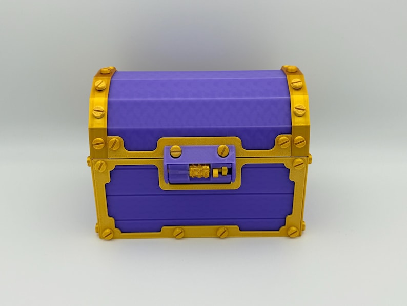 May include: A purple treasure chest with gold-colored accents. The chest features a rectangular shape with a curved top, gold hinges, and a gold clasp. The chest is made of a ridged material and has a lock on the front.