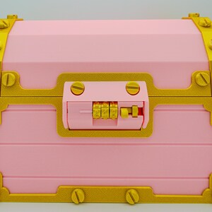 May include: A pink treasure chest with gold-colored trim and hardware. The chest features a combination lock and decorative screw details. The design is reminiscent of a classic pirate chest.