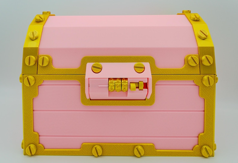 May include: A pink treasure chest with gold-colored trim and hardware. The chest features a combination lock and decorative screw details. The design is reminiscent of a classic pirate chest.