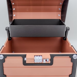 May include: Open, empty treasure chest with a copper-colored exterior and a black interior tray. The chest has a combination lock and is detailed with black trim and silver-colored screws. The handle is made of copper-colored material.
