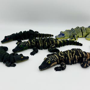 May include: A group of toy alligators in different colours and patterns. The alligators are in shades of green, black, and brown, with detailed textures and realistic features. Some have yellow, green, or red eyes.