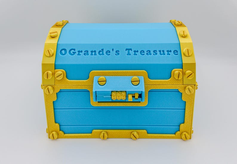 May include: A blue treasure chest with gold-colored accents and the text "OGrande's Treasure" on the lid. The chest has a combination lock and decorative screws. The chest is a decorative storage container.