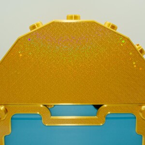 May include: A close-up of a decorative treasure chest. The chest features a gold exterior with a textured surface and a blue interior. Gold accents include screws and decorative trim.