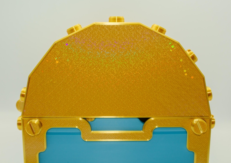 May include: A close-up of a decorative treasure chest. The chest features a gold exterior with a textured surface and a blue interior. Gold accents include screws and decorative trim.