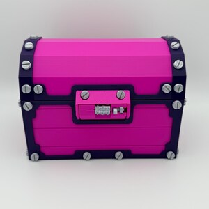 May include: A vibrant pink treasure chest with dark blue trim and silver-colored hardware. The chest features a combination lock and is secured with multiple silver-colored screws. The design is geometric and modern, suitable for storage or decoration.