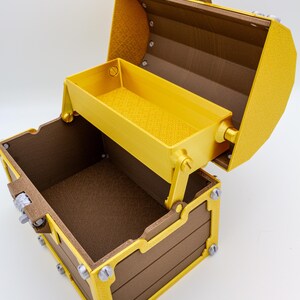 May include: A decorative treasure chest with a hinged lid and a pull-out tray. The chest is brown with gold trim and silver-colored accents. The interior and tray are gold. The chest is open, revealing the interior.