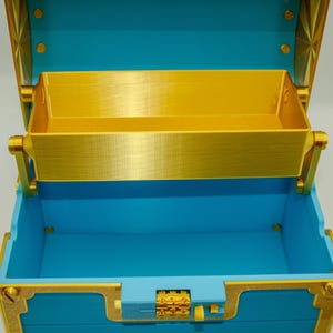 May include: An open, decorative treasure chest with a bright blue exterior and gold-colored accents. The interior features a removable tray. The chest is designed with ornate details, including a decorative clasp and hinges.