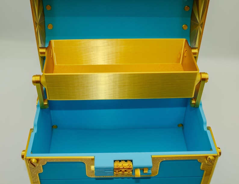 May include: An open, decorative treasure chest with a bright blue exterior and gold-colored accents. The interior features a removable tray. The chest is designed with ornate details, including a decorative clasp and hinges.