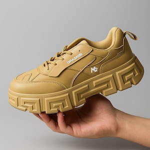 Könnte beinhalten: Beigefarbener Sneaker mit einer dicken, geometrisch gemusterten Sohle. Der Schuh trägt den Markennamen "ORAGAIIBI" und die Initialen "MZ" in weißer Schrift. Der Sneaker wird von einer Hand gehalten.