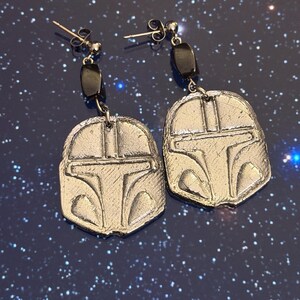 May include: A pair of silver-coloured earrings with a Mandalorian helmet design. Each earring has a post, a small black bead, and a detailed helmet pendant. The earrings are displayed against a dark blue background with white speckles.