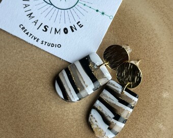 May include: A pair of handmade earrings with a unique design. The earrings feature a black, white, and gold abstract pattern. The earrings are paired with a gold disc and a white card with the text "NAIMA SIMONE CREATIVE STUDIO".