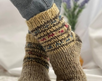 May include: A pair of hand-knitted wool socks. The socks are beige with a patterned cuff featuring brown, blue, and pink accents. The foot of the sock is a darker shade of beige. The socks are worn with blue jeans.