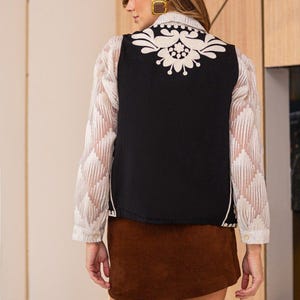 May include: A black sleeveless vest with a white floral embroidery design on the back. The vest is worn over a white sheer blouse and a brown corduroy skirt.