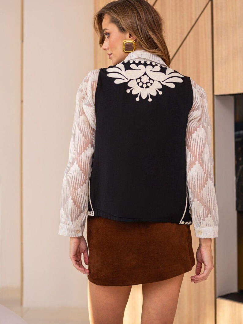 May include: A black sleeveless vest with a white floral embroidery design on the back. The vest is worn over a white sheer blouse and a brown corduroy skirt.