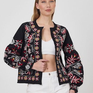 Può includere: Giacca nera ricamata con motivi floreali rossi, bianchi e gialli. La giacca ha maniche lunghe e scollatura rotonda. La modella indossa un top bianco e pantaloni bianchi. La giacca è un'elegante opzione di capospalla.