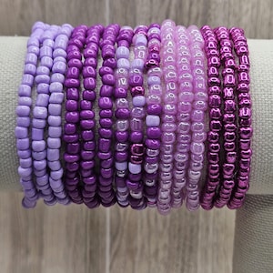 May include: A selection of beaded bracelets in shades of purple, ranging from pale lavender to deep magenta. The bracelets are strung together, displaying the different colours and textures of the beads. The bracelets are displayed on a neutral-coloured surface.