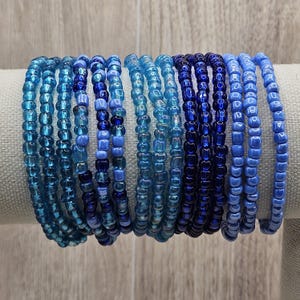 May include: A collection of beaded bracelets in shades of blue, ranging from light turquoise to deep navy. The bracelets are made of small, round beads strung together, creating a stackable accessory. The bracelets are displayed on a neutral-colored surface.