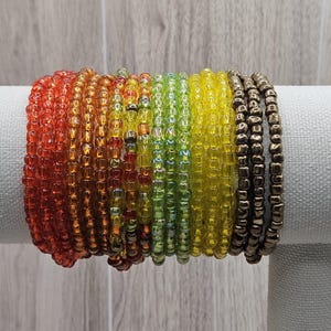 May include: A collection of beaded bracelets in a rainbow of colors, including red, orange, yellow, green, and bronze. The bracelets are made of small, round beads and are stacked together. The bracelets are displayed on a white surface.