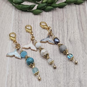 May include: Three decorative charms with gold-colored clasps and whale tail pendants. Each charm features a unique combination of beads in shades of blue, white, gray, and gold. The charms are designed for jewelry or craft projects.
