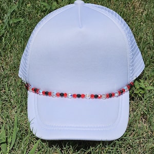 May include: White trucker hat with a mesh back and a curved brim. The brim is adorned with a beaded strand of red, black, and clear beads. The hat is sitting on green grass.