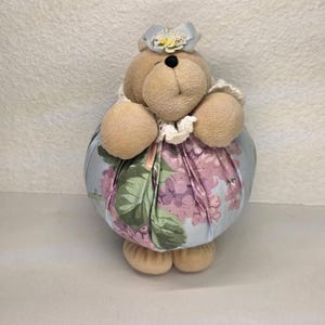 May include: A fabric doorstop in the shape of a bear. The bear has a beige head, arms, and feet, with a black nose and a blue bow with a yellow flower. The body is a round shape covered in a light blue fabric with a floral pattern in pink and green.